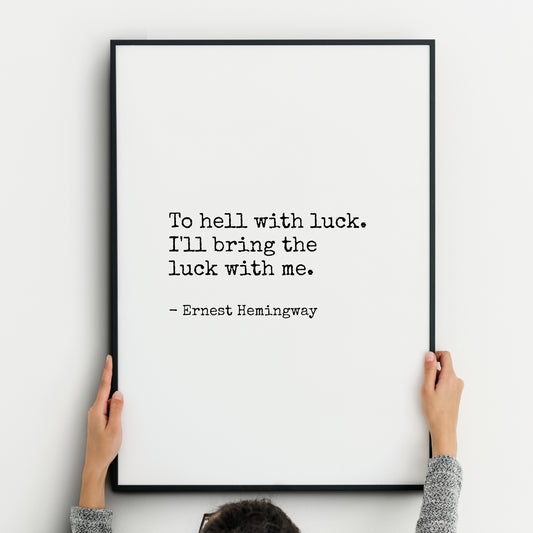 Single portrait typography print featuring an Ernest Hemingway quote in a vintage typewriter font that reads, “To hell with luck. I’ll bring the luck with me.” Text is black on a white background and left-aligned. Styling is minimalist.