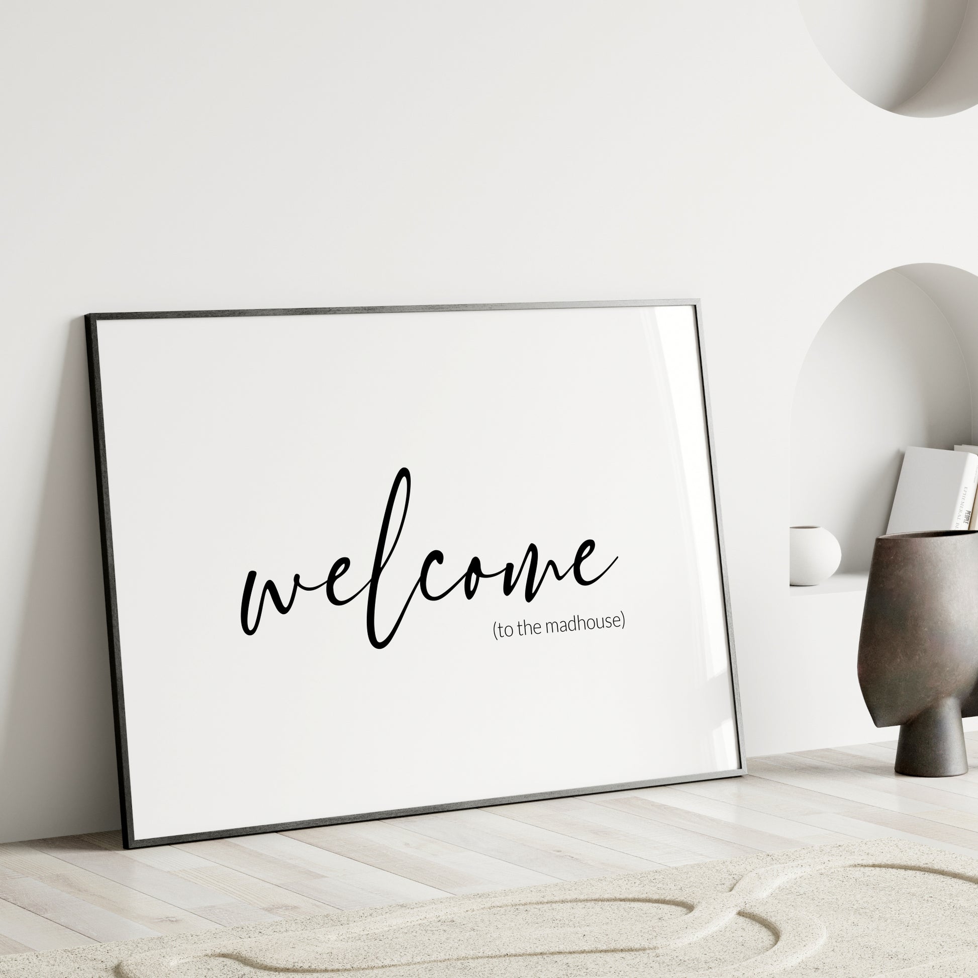 Single landscape typography print. Text reads: welcome (to the madhouse). The word welcome is positioned prominently in a modern script font. Remaining text is in a small, lightweight sans serif font, aligned to the bottom-right of the word welcome. All text is lower case, black on a white background. Styling is elegant and minimalist.