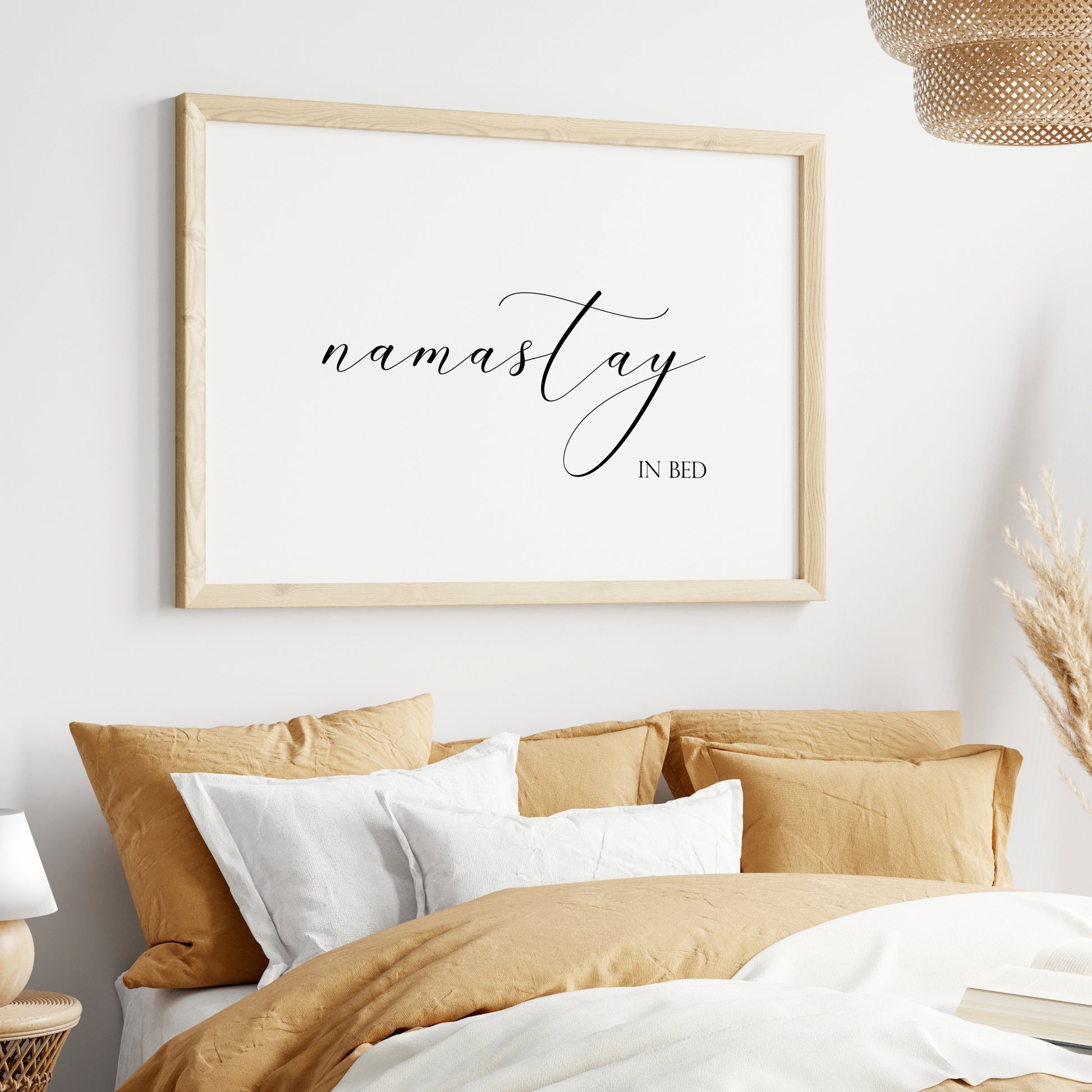 Single landscape typography print. Text reads: namastay in bed. The word namastay is positioned prominently in an elegant script font, with the remaining text in a small upper case serif font aligned to its bottom right. Text is black on a white background.