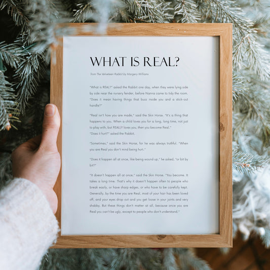Single portrait literary wall art print featuring the quote “What Is Real?” from The Velveteen Rabbit by Margery Williams. The text is left-aligned with elegant serif typography for the title and a fine, modern sans-serif font for the attribution and body text. Printed in black on a clean white background, the design has generous spacing and a calm, balanced layout. Styling is modern and minimalist.